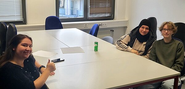 Erasmus-Speed-Dating Drei Schülerinnen sitzen gemeinsam am großen weißen Tisch, lächeln und posieren für ein Foto. Eine Schülerin zeigt den Daumen nach oben.