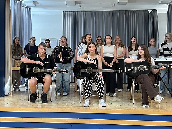 Auf einer Bühne steht eine Gruppe lächelnder junger Menschen. In der hinteren Reihe werden ein Keyboard und ein Xylophon gespielt. Vorne sitzen zwei junge Frauen und ein junger Mann mit Gitarren.