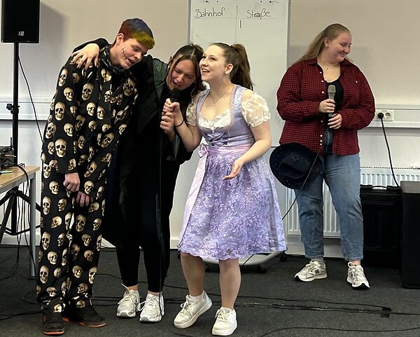 Drei Personen singen zusammen in ein Mikrofon, eine Person in einem lilafarbenen Kleid, eine in einem schwarzen Umhang und eine weitere in einem schwarzen Pyjama mit Totenköpfen. Eine vierte Person im roten Hemd steht daneben mit einem Mikrofon in der Hand. Im Hintergrund ist eine weiße Tafel mit den Wörtern "Bahnhof" und "Straße" zu sehen.