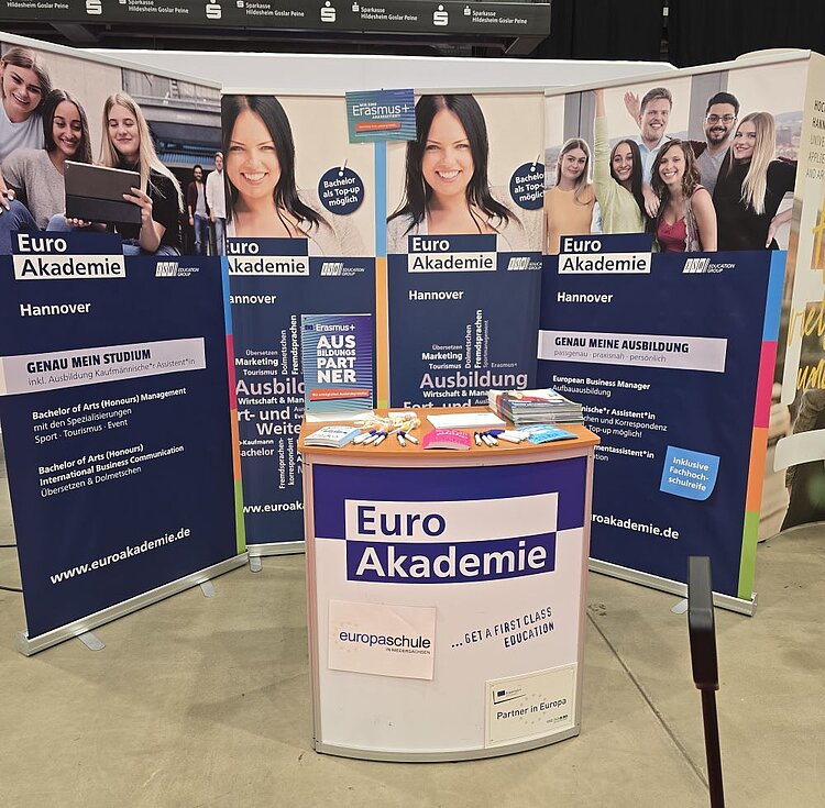 Ein Messestand der Euro Akademie Hannover. Im Vordergrund steht ein weißer Thekentisch mit dem Logo „Euro Akademie“ und dem Slogan „… GET A FIRST CLASS EDUCATION“. Auf dem Tisch liegen Informationsbroschüren, Stifte, ein Notizblock und Lutscher. Im Hintergrund sind mehrere Roll-up-Banner mit Informationen zu Studiengängen wie „Bachelor of Arts (Honours) Management“, „Ausbildung“ und „Erasmus+“ zu sehen. Die Banner zeigen auch Fotos von lächelnden Studierenden und Werbeslogans wie „GENAU MEIN STUDIUM“ und „GENAU MEINE AUSBILDUNG“.