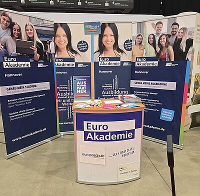 Ein Messestand der Euro Akademie Hannover. Im Vordergrund steht ein weißer Thekentisch mit dem Logo „Euro Akademie“ und dem Slogan „… GET A FIRST CLASS EDUCATION“. Auf dem Tisch liegen Informationsbroschüren, Stifte, ein Notizblock und Lutscher. Im Hintergrund sind mehrere Roll-up-Banner mit Informationen zu Studiengängen wie „Bachelor of Arts (Honours) Management“, „Ausbildung“ und „Erasmus+“ zu sehen. Die Banner zeigen auch Fotos von lächelnden Studierenden und Werbeslogans wie „GENAU MEIN STUDIUM“ und „GENAU MEINE AUSBILDUNG“.
