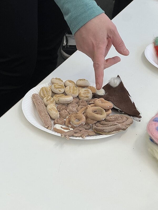 Eine Hand zeigt auf einen Teller mit kunstvollen, essbaren Designs aus Gebäck und Süßigkeiten.