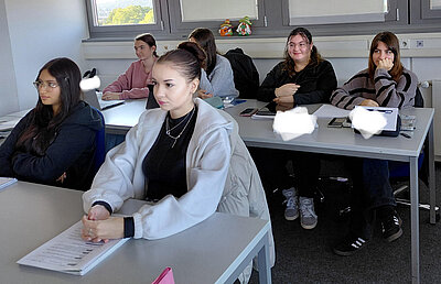 Mehrere Studierende sitzen in Reihen und hören aufmerksam dem Unterricht zu. Auf den Tischen liegen Unterlagen und Laptops, während Sonnenlicht durch die Fenster fällt und den Raum freundlich erhellt.