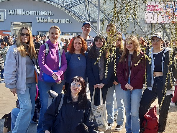 Buchmesse Leipzig: Wo Worte Bildung und Austausch fördern Die Schülergruppe posiert vor dem Messegebäude für ein Gruppenfoto. Herabhängende Äste verdecken einen Teil der Personen. Auf dem Gebäude im Hintergrund steht in großen Lettern "Willkommen. Welcome."