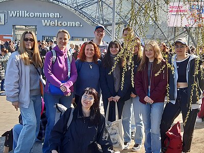 Die Schülergruppe posiert vor dem Messegebäude für ein Gruppenfoto. Herabhängende Äste verdecken einen Teil der Personen. Auf dem Gebäude im Hintergrund steht in großen Lettern "Willkommen. Welcome."