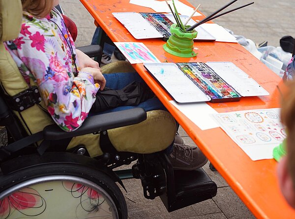 Eine Person im Rollstuhl sitzt an einem Kreativtisch mit Wasserfarben, Pinseln und bemalten Papierblättern.