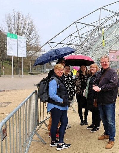 Vor einem großen, gewölbten Glas- und Stahlgebäude vor der Leipziger Buchmesse stehen mehrere Menschen auf sandigem Boden. Einige halten Regenschirme, andere tragen Jacken und Taschen. Im Hintergrund sind Absperrgitter und Hinweistafeln zu sehen. Die Szene wirkt wie ein Eingangsbereich zu einer Veranstaltung. Die Auszubildenden der Euro Akademie posieren mit ihren Lehrkräften für ein Foto.