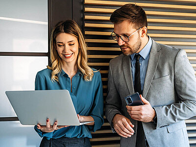 Zwei Personen stehen in einem modernen Büro und arbeiten gemeinsam. Die Frau links trägt eine blaue Bluse und hält einen silbernen Laptop in den Händen. Sie lächelt und wirkt konzentriert. Der Mann rechts trägt einen grauen Anzug, ein blaues Hemd und eine Brille. Er hält ein Smartphone in der Hand und blickt darauf. Im Hintergrund sind eine weiße Tafel und eine Holzvertäfelung zu sehen. Die Atmosphäre ist professionell, kooperativ und zielorientiert.