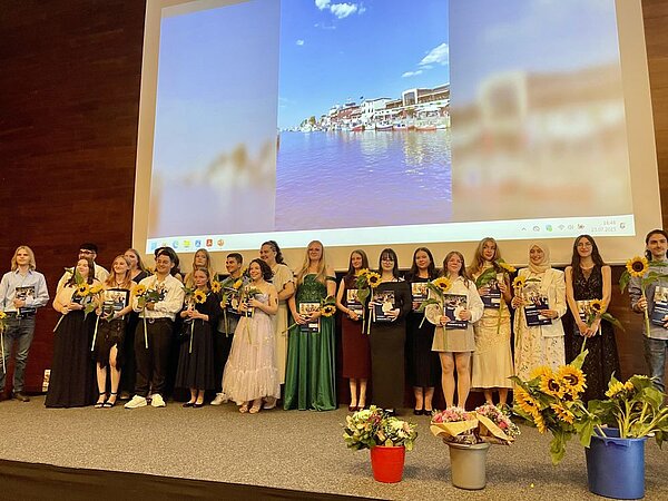 Eine Gruppe von festlich gekleideten Menschen steht auf einer Bühne und hält Zertifikate und Sonnenblumen. Im Hintergrund ist eine Präsentation mit einem Bild einer Stadt am Wasser zu sehen. Auf der Bühne stehen Blumensträuße.