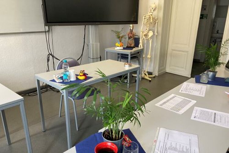 Endspurt für Pflege-Absolvent*innen In einem Raum stehen sich zwei Tische gegenüber. Auf dem rechten befinden sich verschiedenen Blätter mit Tabellen, Zimmerpflanzen in Töpfen, auf dem Tisch gegenüber eine kleine Wasserflasche und Plastikbecher. Hinter dem linken Tisch steht ein Stuhl. An der Wand befindet sich ein großer Bildschirm, daneben ein Skelett. Die Tür des Raums ist zur Hälfte geöffnet.