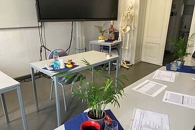 In einem Raum stehen sich zwei Tische gegenüber. Auf dem rechten befinden sich verschiedenen Blätter mit Tabellen, Zimmerpflanzen in Töpfen, auf dem Tisch gegenüber eine kleine Wasserflasche und Plastikbecher. Hinter dem linken Tisch steht ein Stuhl. An der Wand befindet sich ein großer Bildschirm, daneben ein Skelett. Die Tür des Raums ist zur Hälfte geöffnet.