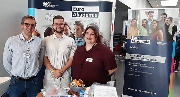Euro Akademie Mainz auf BIM Rheinhessen Drei Personen stehen lächelnd hinter dem Messestand der Euro Akademie Mainz. Auf dem Tisch liegen Broschüren, eine Schüssel mit Süßigkeiten und Stifte, während im Hintergrund ein Roll-Up mit dem Logo der Euro Akademie zu sehen ist.