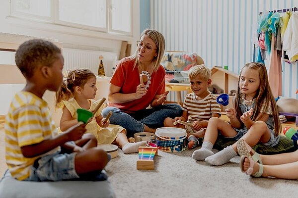 Eine Gruppe von Kindern sitzt mit einer weiblichen Erzieherin im Kreis auf einem hellen Teppichboden. Die Kinder halten bunte Musikinstrumente wie Rasseln, Xylophon und Tamburin in den Händen. Im Hintergrund stehen ein Regal mit Spielzeug und eine Kleiderstange mit bunter Kleidung. Tageslicht fällt durch ein großes Fenster in den Raum.