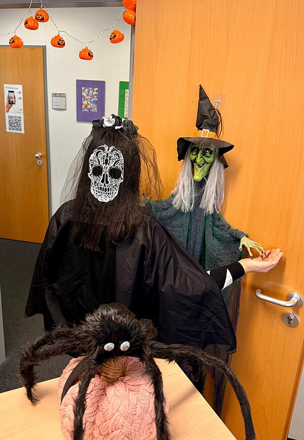 Gruselstimmung pur an der Euro-Schule & Euro-Akademie Dresden! Im Vordergrund sitzt die gleiche schwarze Spinne auf dem rosafarbenen Kürbis, im Hintergrund sind eine Person mit Totenkopfmaske und eine Hexendekoration an der Tür zu sehen.