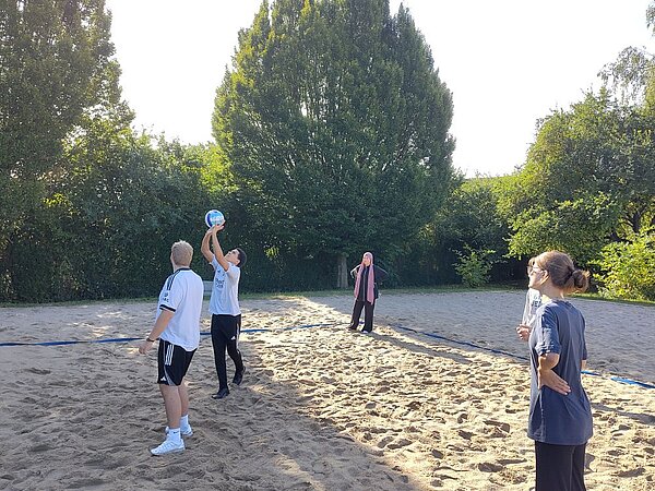 Mehrere Personen spielen auf einem Sandplatz Beachvolleyball. Eine Person streckt seine Arme nach oben, um den Ball zu stoßen.