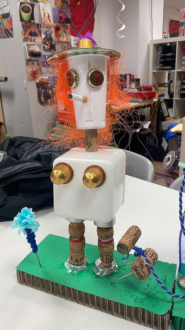 Eine Figur bestehend aus einem viereckigen Plastikbauch, einem Plastikbecher als Kopf, mit Korken als Füßen und Kronkorken als Augen und Brust, steht auf grüner Pappe.