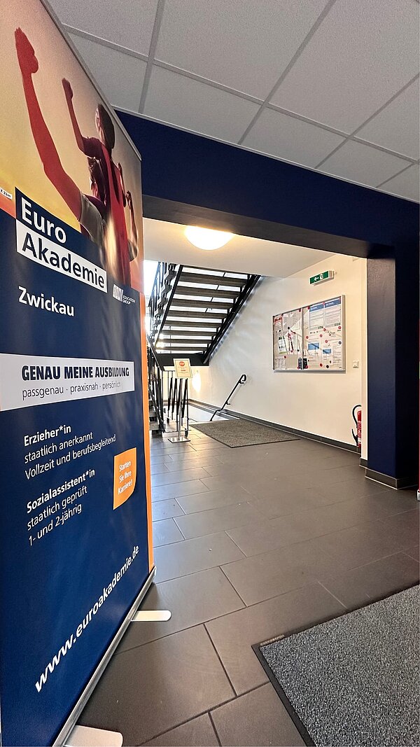 Aus Rot wird Blau: Malerarbeiten abschlossen Links im Bild steht ein Roll-up der Euro Akademie, dass die angebotenen Ausbildungen bewirbt. Das restliche Bild zeigt den grau gefliesten Eingangsbereich der Euro Akademie Zwickau mit dem neu gestrichenen Durchgang zum Treppenhaus.