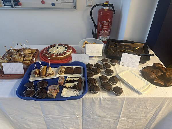 Ein Tisch mit selbstgebackenem Kuchen und Gebäck, beschriftet mit Preisschildern.