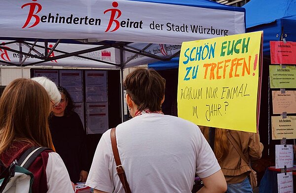 Besucher*innen stehen am Infostand des Behindertenbeirats der Stadt Würzburg; ein großes Schild trägt die Aufschrift „Schön euch zu treffen! Warum nur einmal im Jahr?“.