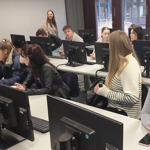 In einem modernen Computerraum der Universitätsbibliothek Würzburg sitzen mehrere angehende Erzieher der Euro Akademie Tauberbischofsheim an langen Tischen mit Desktop-Computern. Die Personen sind in lockerer Kleidung, darunter eine Frau in einem weiß-schwarzen gestreiften Pullover, die nach rechts blickt. Im Hintergrund sind große Fenster mit Blick auf eine Außenfassade zu sehen, die Tageslicht in den Raum lassen. Die Atmosphäre wirkt konzentriert und lernorientiert, während die Gruppe an der Exkursion teilnimmt.