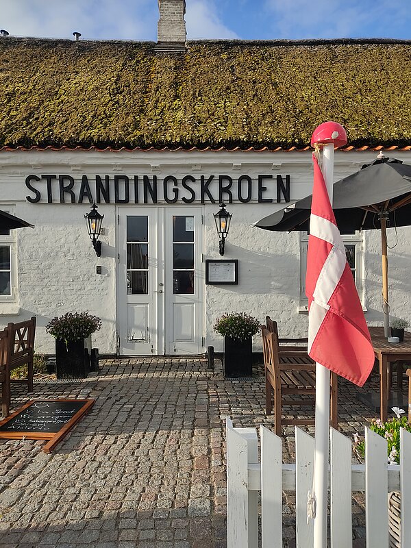 Erfahrungsbericht Erasmus – Hygge von Janice Weßel Ein traditionelles dänisches Gasthaus mit weißer Fassade und einem mit Moos bewachsenen Reetdach. Über der Tür hängt ein Schild mit der Aufschrift „STRANDINGSKROEN“. Vor dem Eingang stehen Holztische, Blumen und eine dänische Flagge. Die gepflasterte Terrasse vermittelt eine gemütliche Atmosphäre.