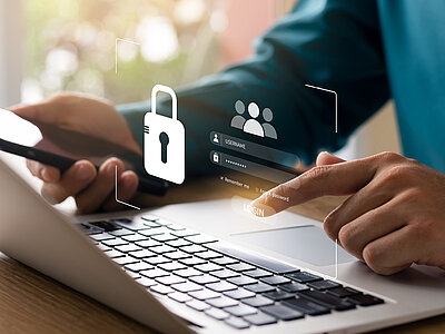 Eine Person arbeitet an einem Laptop und hält gleichzeitig ein Smartphone in der Hand. Auf dem Laptop-Bildschirm ist ein Login-Fenster zu sehen, das Benutzername und Passwort abfragt. Über dem Laptop schwebt ein Symbol eines Vorhängeschlosses, das Sicherheit oder Datenschutz symbolisiert, sowie ein Benutzersymbol, das auf den Login-Prozess hinweist.
