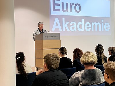 Ein Mann hält am Rednerpult der Georg-Friedrich-Händel-Halle eine Ansprache; hinter ihm wird das Logo der Euro Akademie auf eine große Leinwand projiziert.