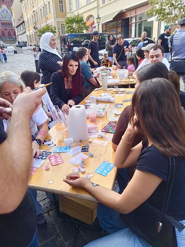 Lange Holztafel im Freien, an der mehrere Menschen unterschiedlichen Alters sitzen. Auf dem Tisch liegen Bastelmaterialien, bunte Farbtöpfchen, Pinsel, Papierrollen und kleine Gegenstände. Einige Personen sind in kreative Tätigkeiten vertieft, andere unterhalten sich. Im Hintergrund sind Kopfsteinpflaster, historische Gebäude und weitere Passanten zu sehen. Die Szene wirkt lebendig und gemeinschaftlich.