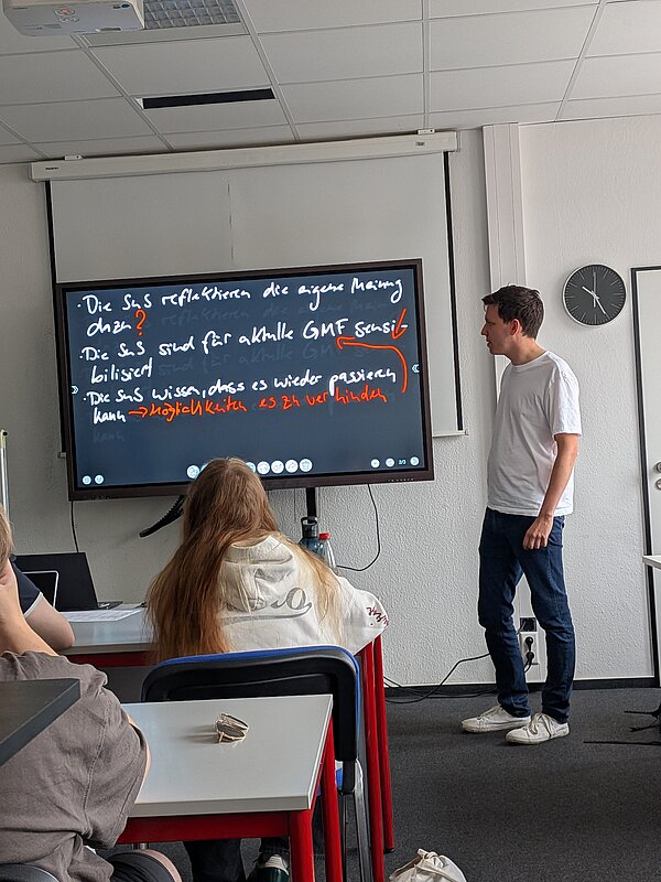 Ein Schüler präsentiert vor der Klasse an einem digitalen Whiteboard. Darauf sind handschriftliche Stichpunkte und Anmerkungen zu einem Unterrichtsthema zu sehen.