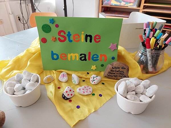 Auf einem Tisch liegen weiße Steine, bunte Stifte in einem Becher stehen bereit zum Steinemalen, wie die bunte Karte verrät.