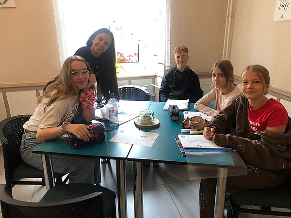 Sprachprojekte mit internationalem Flair Vier Mädchen und ein Junge im Teenageralter sitzen an einem dunkelgrünen Tisch und schreiben auf weißen Zetteln Notizen auf, dabei schauen sie Richtung Kamera.