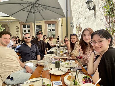 Eine Gruppe junger Erwachsener sitzen im Außenbereich eines Restaurants an einem Tisch und lächeln. Auf dem Tisch stehen leere Teller und Gläser. Im Hintergrund sind ein altes Gebäude und ein großer Sonnenschirm zu sehen.