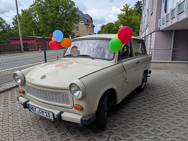 Ein cremefarbener Trabant mit eckiger Karosserie steht auf einem gepflasterten Parkplatz vor einem Wohnhaus. An den Außenspiegeln und der Antenne sind bunte Luftballons befestigt. Die Motorhaube zeigt einen großen, aufgeklebten roten Kreis. Die Fenster sind heruntergekurbelt, das Lenkrad und die Armaturen sind sichtbar. Im Hintergrund stehen Bäume und weitere Gebäude.