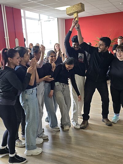 Die gut gelaunten weiblichen und männlichen Auszubildenden stehen beisammen und halten siegessicher einen großen Holzhammer in die Lüfte.