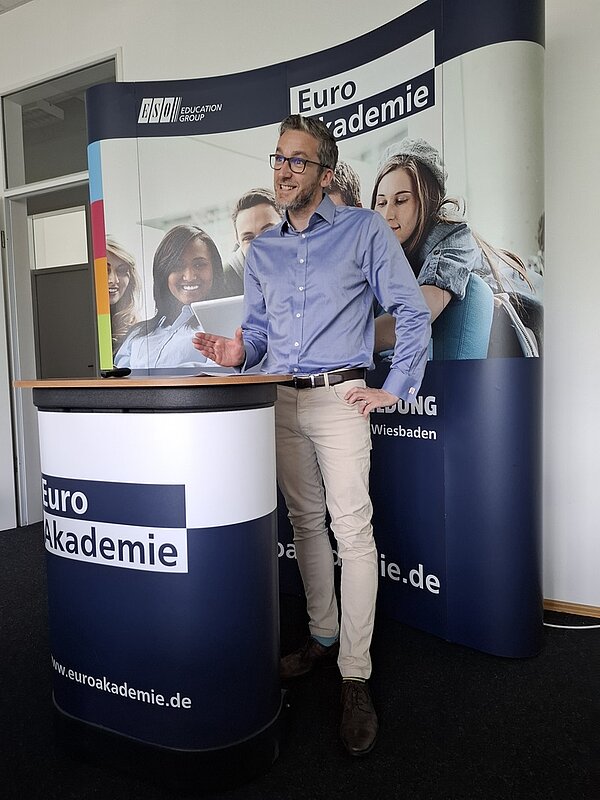 Wir sind Fremdsprachenkorrespondent*innen! Englischlehrer Lee Hawkins steht gut gelaunt an einem Euro Akademie-Redepult und spricht zu den Absolventinnen und Absolventen.