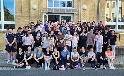 Eine große Gruppe von etwa 80 Personen posiert für ein gemeinsames Foto vor einem gelben Backsteingebäude. Viele lächeln, einige machen Handzeichen.