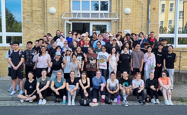 Eine große Gruppe von etwa 80 Personen posiert für ein gemeinsames Foto vor einem gelben Backsteingebäude. Viele lächeln, einige machen Handzeichen.