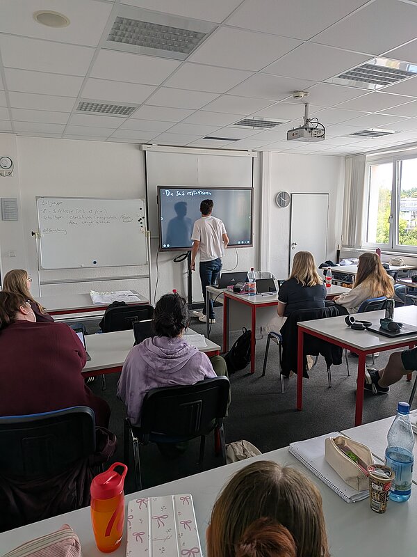 Unterrichtssituation in einem Klassenraum: Ein Schüler steht vorne an einem digitalen Whiteboard und arbeitet daran, während die übrigen Schüler*innen an ihren Tischen sitzen und zuhören.
