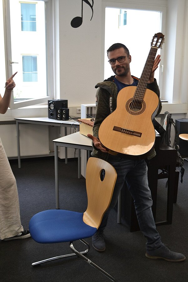 Ein Auszubildender der Euro Akademie Würzburg hält lächelnd eine holzerne Akustikgitarre in die Kamera. 