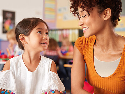 Auf dem Bild sind zwei Personen im Klassenzimmer zu sehen. Die Person links trägt eine weiße Bluse mit bunten Pompons an den Ärmeln, während die Person rechts ein orangefarbenes Oberteil und lockiges Haar hat. Im Hintergrund sind Kinderzeichnungen, pädagogische Poster und andere Lernmaterialien an den Wänden zu erkennen.