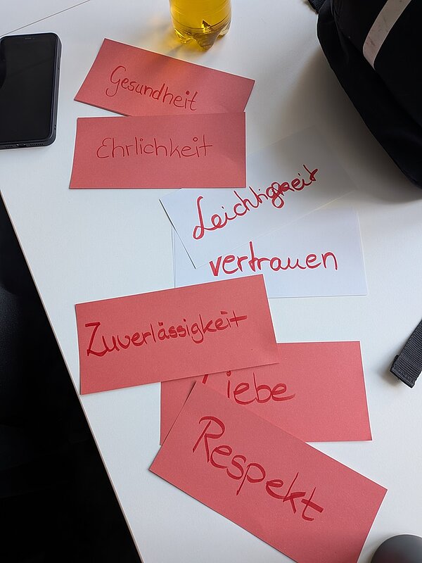 Mehrere beschriftete Karten mit handschriftlichen Begriffen wie „Gesundheit“, „Ehrlichkeit“, „Leichtigkeit“, „Vertrauen“, „Zuverlässigkeit“, „Liebe“ und „Respekt“ liegen auf einem Tisch.