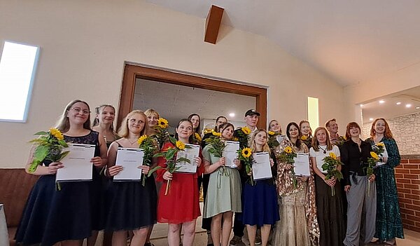 Feierliche Übergabe der Zeugnisse In einem Raum halten Schülerinnen und Schüler in festlicher Kleidung ihre Zeugnisse in die Kamera. Außerdem haben alle noch eine kleine Sonnenblume in der Hand.