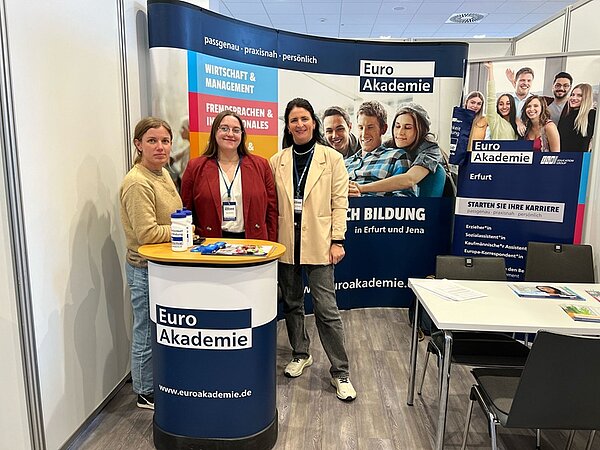 Gelungene Messeteilnahme Drei Mitarbeiterinnen stehen gut gelaunt an einem Stehtisch am Messestand der Euro Akademie, hinter ihnen ein großes Roll-Up und ein Tisch mit vier Stühlen.