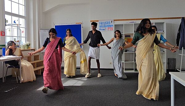 Eine Gruppe indischer Auszubildender in farbenfrohen Saris und traditioneller Kleidung tanzt mit ausgebreiteten Armen in einem Klassenraum, während eine Frau am Tisch zusieht.
