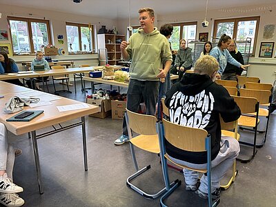  Ein Mann in einem hellgrünen Kapuzenpulli erklärt oder diskutiert mit anderen Personen in einem hellen Klassenraum, während einige sitzen und zuhören. Im Hintergrund sind weitere Tische mit Materialien und Bastelsachen zu sehen. Die Atmosphäre wirkt lebendig und interaktiv, wie bei einer Gruppenarbeit oder einem Workshop.