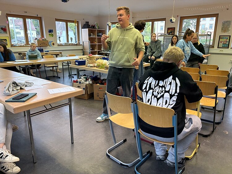 Neues Kreativprojekt: Komm, wir bauen eine Stadt! Ein Mann in einem hellgrünen Kapuzenpulli erklärt oder diskutiert mit anderen Personen in einem hellen Klassenraum, während einige sitzen und zuhören. Im Hintergrund sind weitere Tische mit Materialien und Bastelsachen zu sehen. Die Atmosphäre wirkt lebendig und interaktiv, wie bei einer Gruppenarbeit oder einem Workshop.
