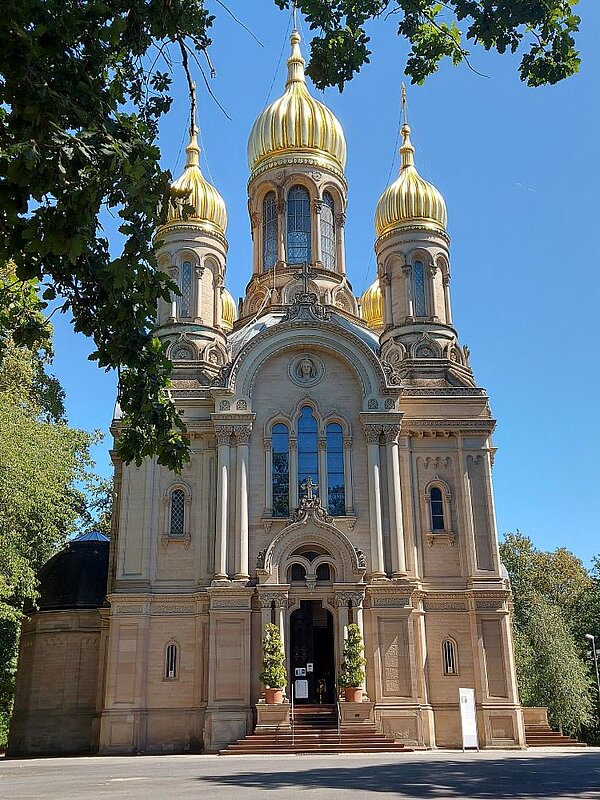 Eine prächtige historische Kirche oder Kathedrale mit goldenen Kuppeln und Säulen, umgeben von grünen Bäumen und einem klaren blauen Himmel.