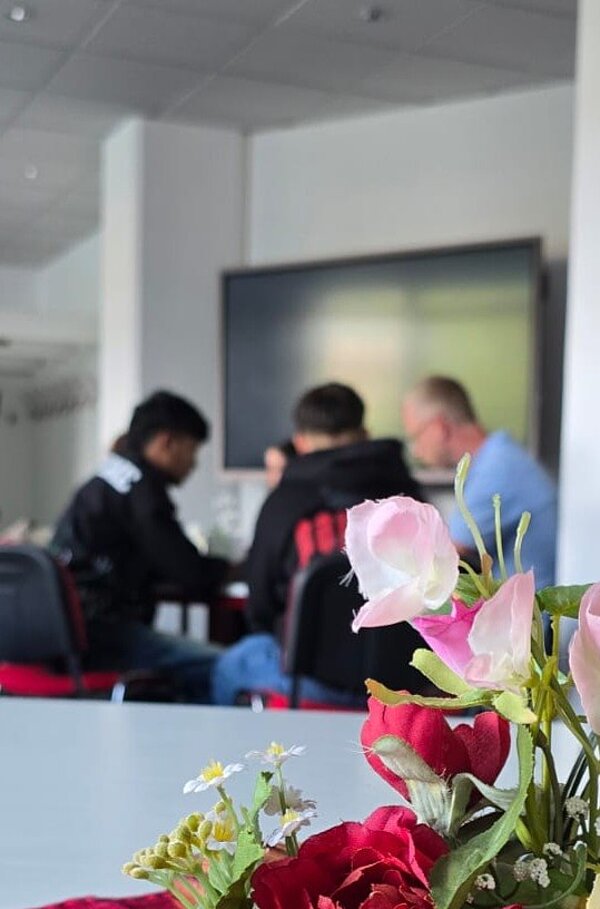 Im Vordergrund stehen rote und rosa künstliche Blumen auf einem weißen Tisch. Im Hintergrund sitzen drei Personen an einem Tisch, die sich miteinander unterhalten. Die Szene spielt in einem hellen Raum mit einem großen Bildschirm an der Wand.