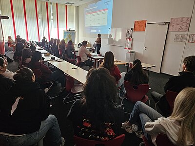 Mehrere Schülerinnen und Schüler sitzen in einem Klassenraum und verfolgen eine Präsentation. Vorne steht eine Referentin, die Folien mit Statistiken und Informationen an eine Leinwand projiziert. Links ist eine Landkarte auf einem Stativ zu sehen, die Tische sind mit Laptops, Notizbüchern und Handys belegt.