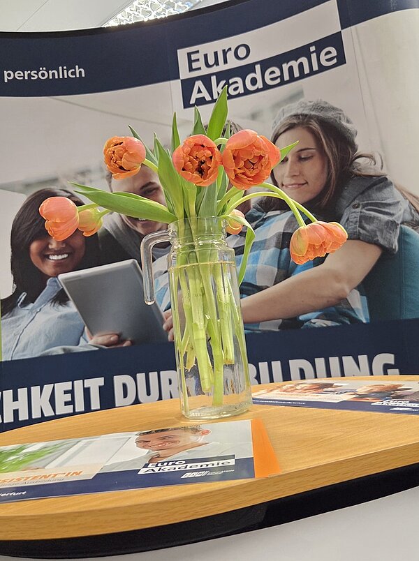 Ein transparenter Glaskrug mit orangefarbenen Tulpen steht auf einem Tisch. Im Hintergrund ist ein Roll-up mit dem Schriftzug 'Euro Akademie' und teilweise verdeckten Gesichtern zu sehen. Auf dem Tisch liegen mehrere Broschüren oder Flyer zur Euro Akademie. Eine einladende Empfangssituation an einem Informationsstand.
